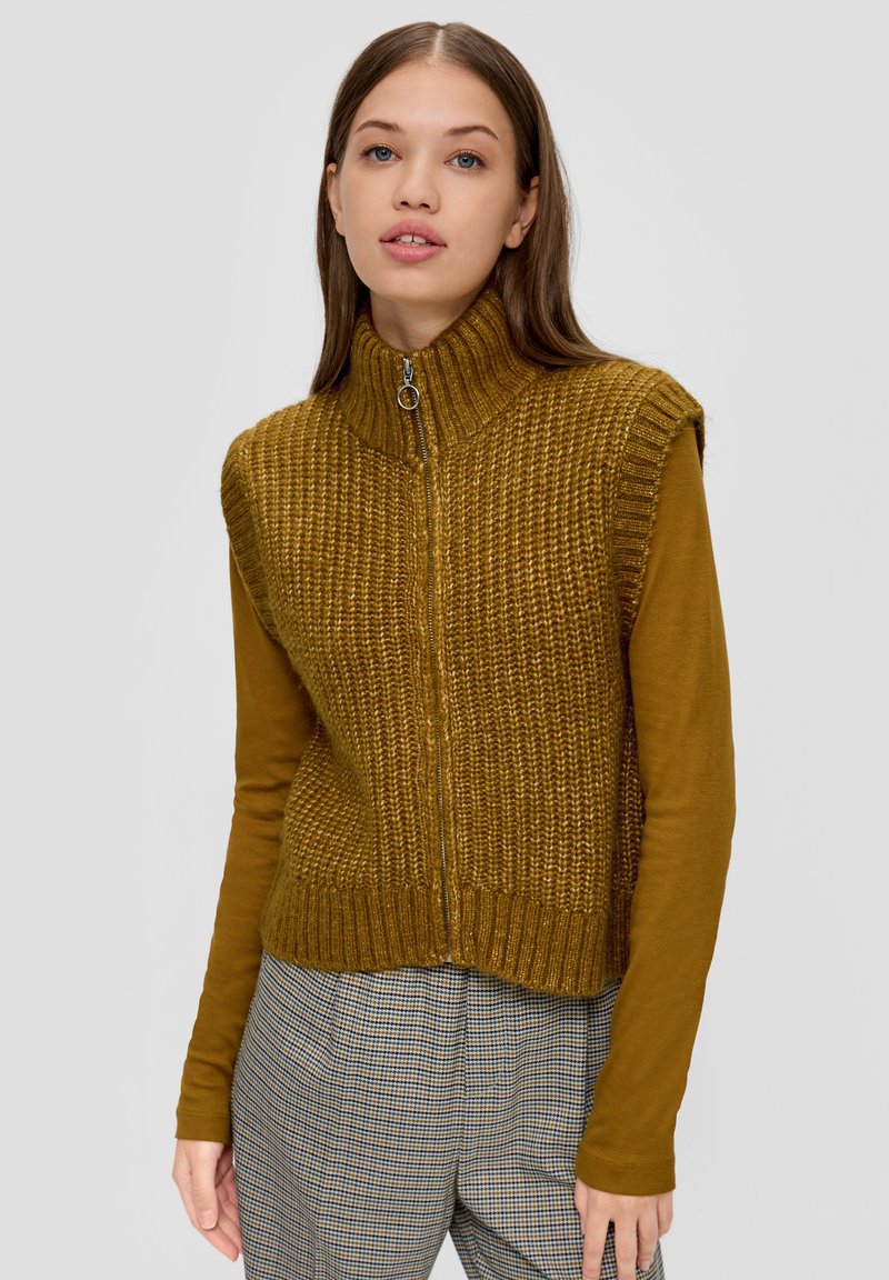 Strickweste in olivgrün mit hohem Kragen und halbem Reißverschluss. Begleitet von einem braunen Langarmshirt und karierten Hosen.