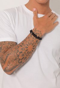 Mustmust ja mitme kihiga musta helmesed, millel on ümmargused ja tekstureeritud helmed, kantud käel, millel on keerukad tätoveeringud, valge T-särgi taustal.