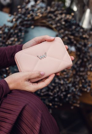 Pink quilted leather wallet featuring a silver logo accent, with a smooth texture and zip closure. Hands holding the wallet are visible.