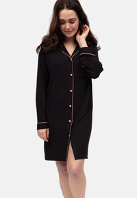 Camisón de algodón negro con ribete rosa, mangas largas, cierre de botones en el frente y una abertura lateral para mayor movimiento. Presenta un bolsillo en el pecho.