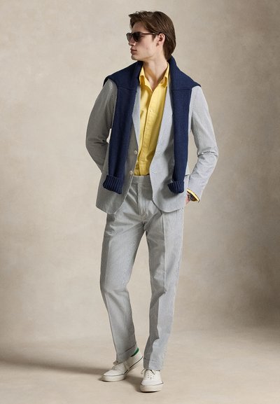 Homme portant un costume à fines rayures, une chemise jaune, un pull bleu marine drapé sur les épaules, des baskets blanches et des lunettes de soleil, debout devant un fond beige.