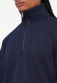 Donker marineblauwe quarter-zip pullover gemaakt van zacht materiaal. Voorzien van een metalen rits en een hoge kraag. Slanke snit, minimalistisch ontwerp zonder zichtbare patronen.