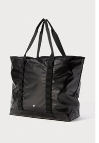 Bolso tote grande negro con dobles asas largas, correas verticales texturizadas y un pequeño logo circular blanco en el centro frontal.