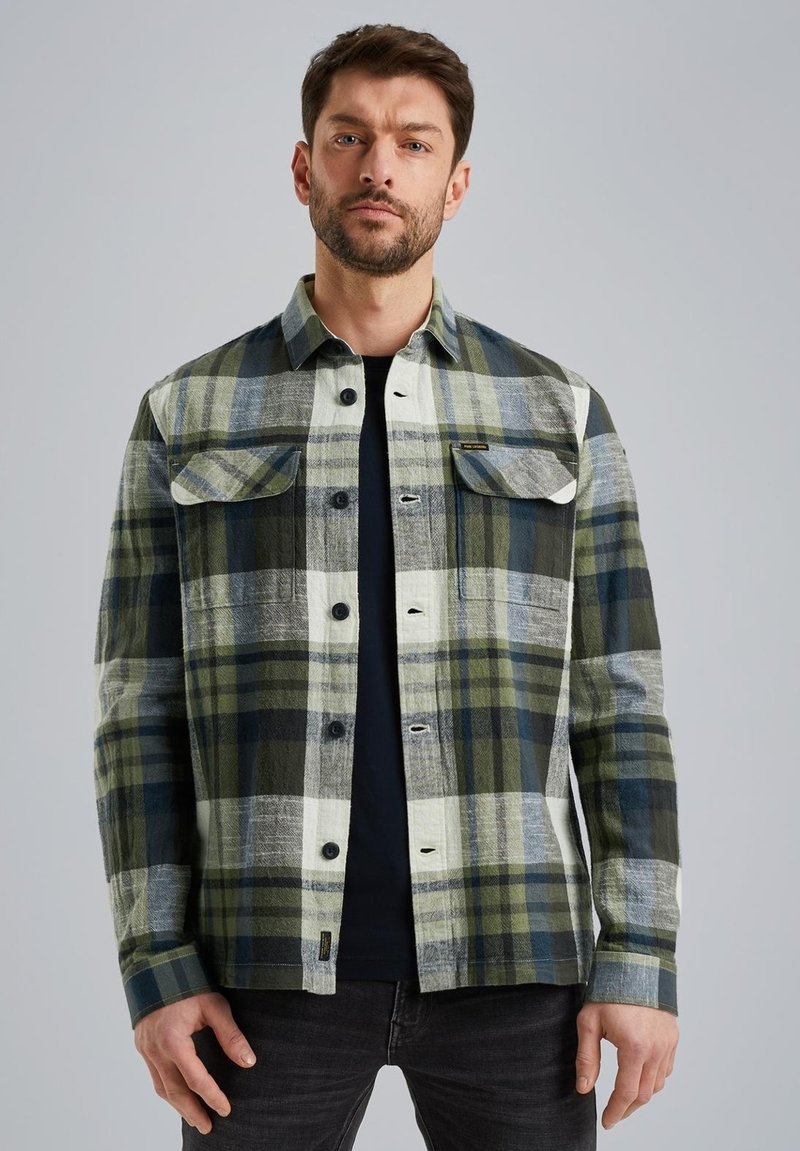 Chemise à carreaux verte et grise avec des manches longues, fermeture à boutons, deux poches poitrine et une coupe décontractée, portée sur un t-shirt noir.