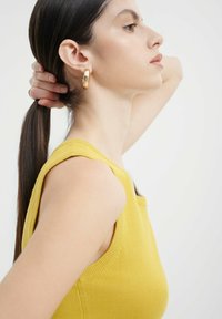 Gelbes ärmelloses Oberteil mit gerippten Kanten, kombiniert mit goldenen Creolen. Die Haare zu einem glatten Pferdeschwanz gestylt, was das Seitenprofil betont.
