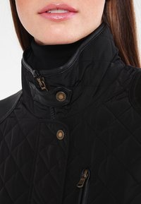 Veste matelassée noire avec un col haut, dotée d'accents en cuir et de boutons en laiton. Comprend des poches zippées et une texture lisse.