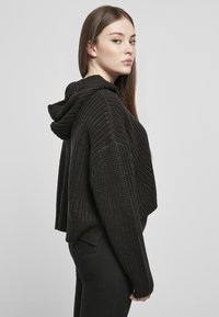 Fekete, rövidített kötött kapucnis pulóver, texturált mintával, széles ujjakra és húzózsinóros kapucnira tervezték. Slim fit fekete nadrág fölött viselik.