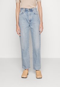 Jeans de tiro alto en azul claro con corte recto, que presentan presillas para el cinturón, diseño de cinco bolsillos y secciones desgastadas. Combinados con zapatillas de lona beige.