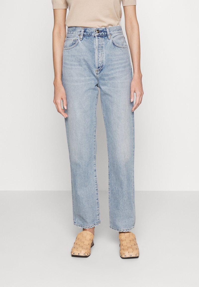 Jeans de tiro alto en azul claro con corte recto, que presentan presillas para el cinturón, diseño de cinco bolsillos y secciones desgastadas. Combinados con zapatillas de lona beige.