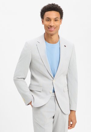 Junger Mann steht, trägt ein hellblaues Hemd und einen weißen Anzug mit dünnen blauen vertikalen Streifen, lächelnd, eine Hand in der Tasche.