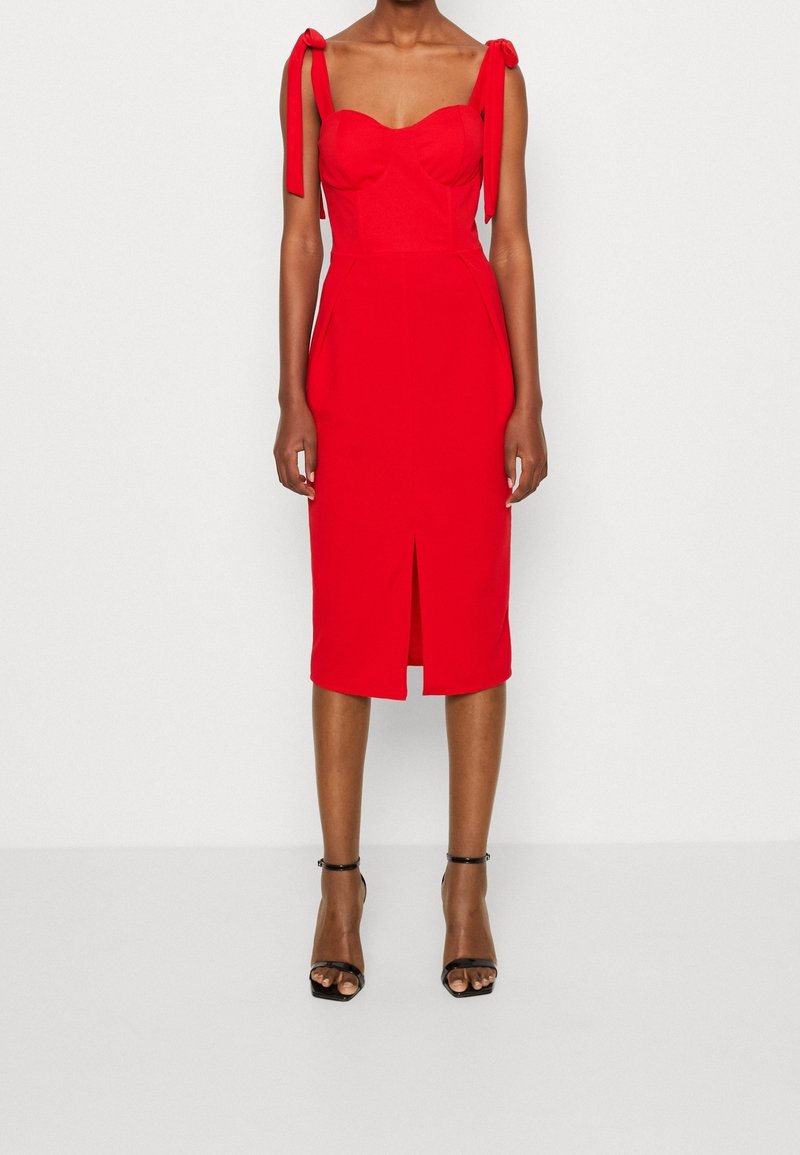 Femme portant une robe midi rouge sans manches avec des bretelles à nouer et une fente devant, associée à des sandales noires à talons hauts avec bride à la cheville.