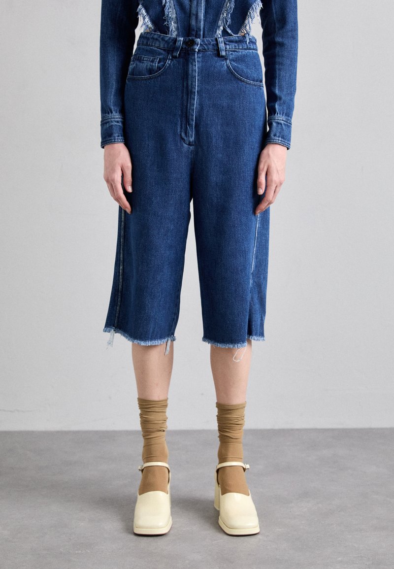 Denim culottes i mellomblått med en frynset kant, med frontlommer og høy midje. Kombinert med beige strømpebukser og kremfargede sko.