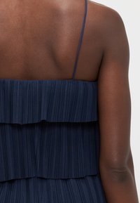 Navy pleated dress with layered ruffles, adjustable spaghetti straps, and a smooth texture, shown from the back.