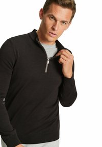 Felpa nera con zip, con orlo e polsini a coste; texture a maglia fine; modello che tiene la zip, indossata sopra una maglietta grigia.