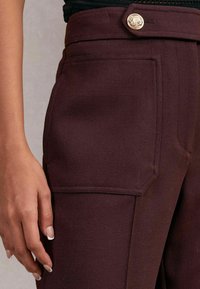 Közelkép egy kézről, amely sötétbarna nadrág mellett pihen, amelynek nagy zsebe van és arany gomb található a derekán.