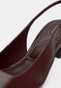 Burgunderfarbener Slingback-Schuh mit glattem Lackfinish, offenen Seiten, niedrigem quadratischem Absatz und geometrischen Details auf der Innensohle.