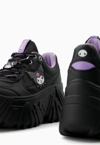 Black sneakers with a chunky sole feature purple accents, a cartoon character emblem, and textured rubber branding on the tongue.
