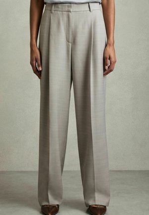 Person trägt lose, hoch taillierte beige Hose und braune, spitze Schuhe, steht vor einem schlichten grauen Hintergrund.