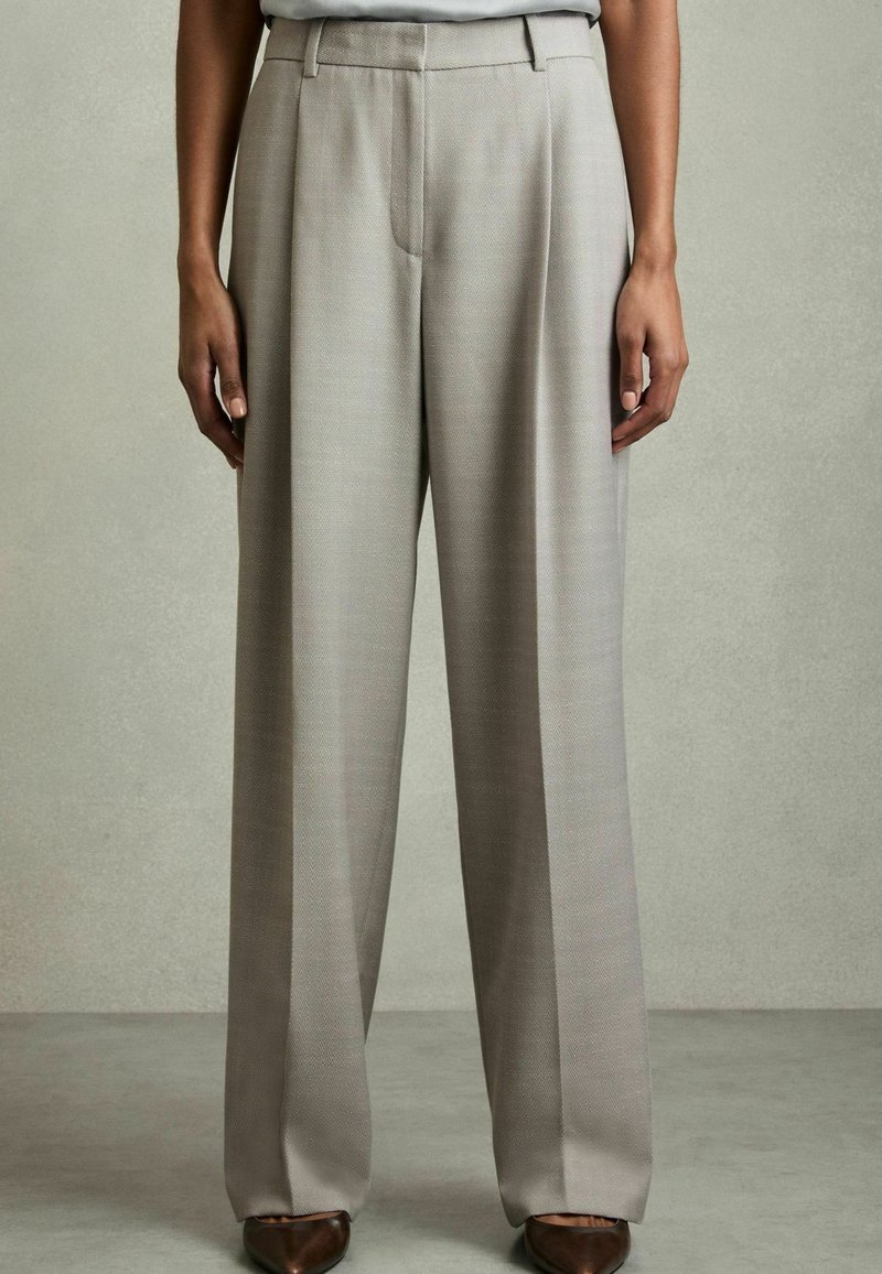 Personne portant un pantalon beige ample et taille haute avec des chaussures brunes pointues, debout devant un fond gris uni.