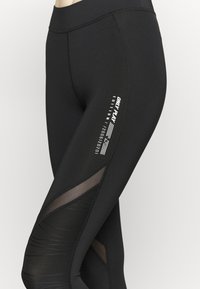 Czarne legginsy sportowe z wysokim stanem, panelem z siateczkowymi wstawkami oraz białymi nadrukowanymi detalami logo po bokach.
