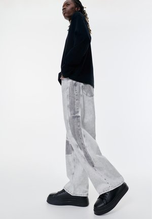 Persoon met gevlochten haar die een zwarte top met lange mouwen draagt, brede acidwash jeans, zwarte platform sneakers, staand tegen een witte achtergrond.