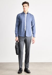 Modrá košeľa na zapínanie s golierom v kombinácii so sivými šitými nohavicami a čiernymi koženými topánkami, drží čiernu koženú tašku. Hladká textúra látky a priliehavý strih.