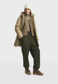 Parka beige con dettagli trapuntati, pantaloni cargo verde oliva, maglione beige e scarponi da trekking marroni. Cappello lavorato a maglia bianco con accentuazione del logo.