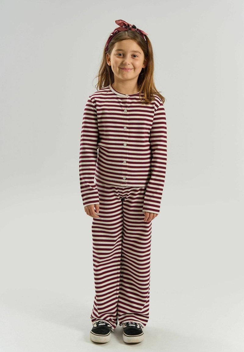 Fille souriante, debout, portant un pyjama rayé horizontal bordeaux et blanc avec un bandeau assorti, baskets noires sur fond uni.