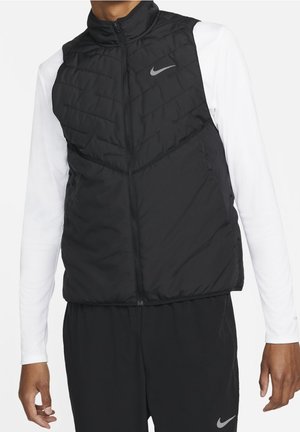 Homme portant une veste sans manches matelassée noire sur une chemise blanche à manches longues et un pantalon noir, tous deux avec logos Nike visibles.