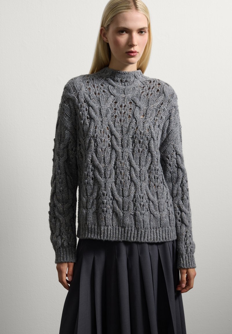 Femme portant un pull en maille torsadée gris avec un col montant et une jupe plissée noire, se tenant devant un fond clair uni.