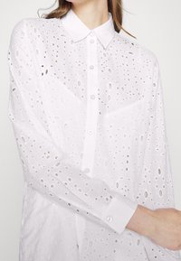 Camisa blanca de botón con intrincados patrones recortados, cuello amplio y mangas largas con puños abotonados. Fabricada en tejido ligero.