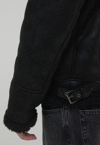 Giubbotto in shearling nero con vita regolabile, maniche texturizzate e polsini pelosi, arricchito da un dettaglio con fibbia metallica.