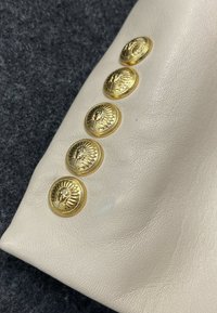 Cinq boutons décoratifs dorés ornés d'un emblème de visage détaillé, disposés en ligne diagonale sur une surface en cuir crème.