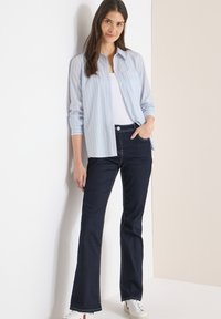 Chemise à rayures bleues et blanches avec boutonnière et poche poitrine, portée sur un débardeur blanc, associée à un jean évasé foncé et des baskets blanches.