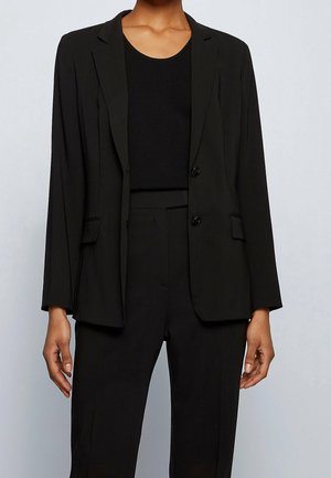 Person iført en sort blazer og matchende sorte bukser over en sort rundhalset top, stående med afslappede hænder mod en ensfarvet baggrund.