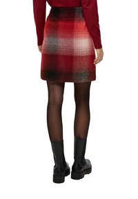 Femme portant une mini-jupe écossaise rouge, des collants noirs transparents, des bottines noires à la cheville et un haut rouge à manches longues, vue de dos.