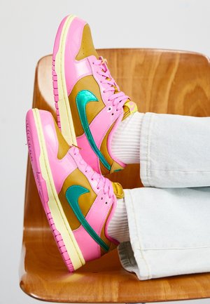 Stopy w różowych, brązowych i turkusowych butach sportowych Nike z białymi skarpetkami i jasnymi dżinsami, spoczywające na drewnianym krześle.