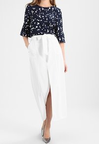 Mujer que lleva una blusa azul marino con lunares blancos y una falda blanca de cintura alta con abertura frontal y cinturón anudado, combinada con tacones plateados.