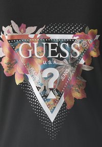 Černá látka s květovaným obráceným trojúhelníkem, stříbrnými nýty a textem "GUESS U.S.A. Washované džíny" s otazníkem.