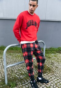 Röd sweatshirt med texten "JORDAN", rutiga svarta och röda byxor och svarta sneakers. Materialet verkar mjukt med ribbade mudd och midjeband.