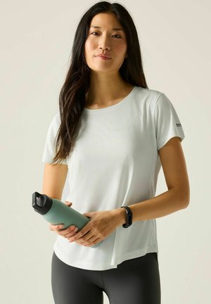 Kobieta w białej koszulce z krótkim rękawem i czarnych legginsach trzyma zieloną butelkę na wodę, na lewym nadgarstku ma czarny fitness tracker.