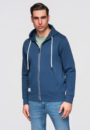 Jonge man die een blauwe hoodie met ritssluiting en witte trekkoorden en lichtblauwe spijkerbroek draagt, staand tegen een effen achtergrond.