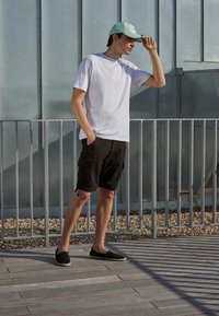 White short-sleeve t-shirt, black cargo shorts with side pockets, light blue cap, and black espadrille shoes on a wooden deck.