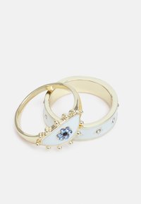 Anéis em tom dourado apresentando um design de olho em esmalte branco com pedras azuis e transparentes, acentos texturizados em ouro e bandas circulares lisas.
