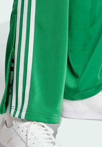 Green athletic pants with white stripes and zipper detail, paired with white shoes featuring textured material and branding accents.