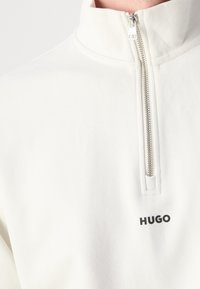 Sweat-shirt blanc à col montant avec fermeture éclair argentée jusqu'au quart, arborant un petit logo noir "HUGO" sur la poitrine.