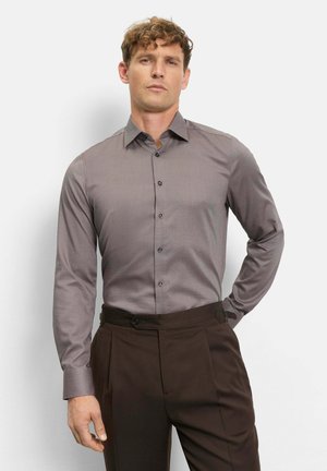 Mann mit lockigem Haar trägt ein tailliertes graues Hemd, das in hoch taillierte, dunkelbraune Hosen gesteckt ist, und steht mit einer Hand hinter dem Rücken.