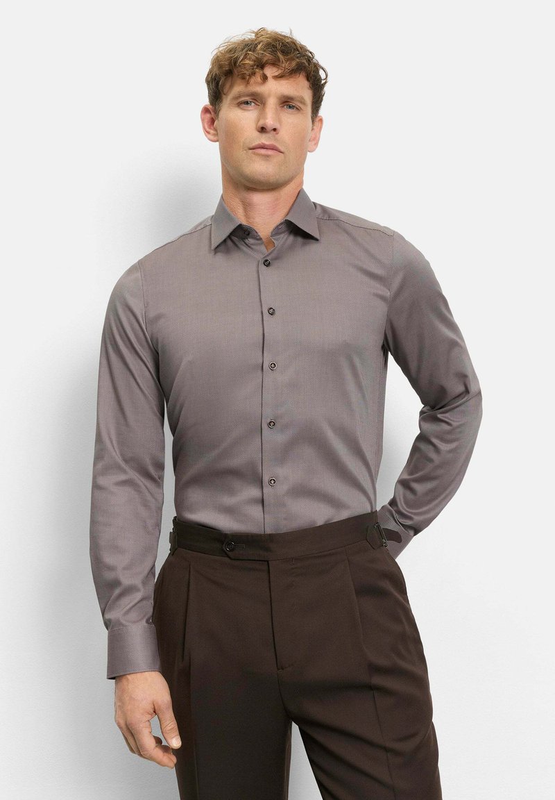 Mann mit lockigem Haar trägt ein tailliertes graues Hemd, das in hoch taillierte, dunkelbraune Hosen gesteckt ist, und steht mit einer Hand hinter dem Rücken.