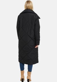 Cappotto lungo trapuntato nero con un grande colletto, caratterizzato da motivi a diamante e maniche lunghe, indossato sopra jeans blu e stivali beige.
