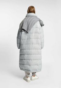 Abrigo puffer gris con un suéter gris de canalé sobre los hombros. Presenta un diseño hasta la rodilla y una textura acolchada. Zapatillas blancas visibles.